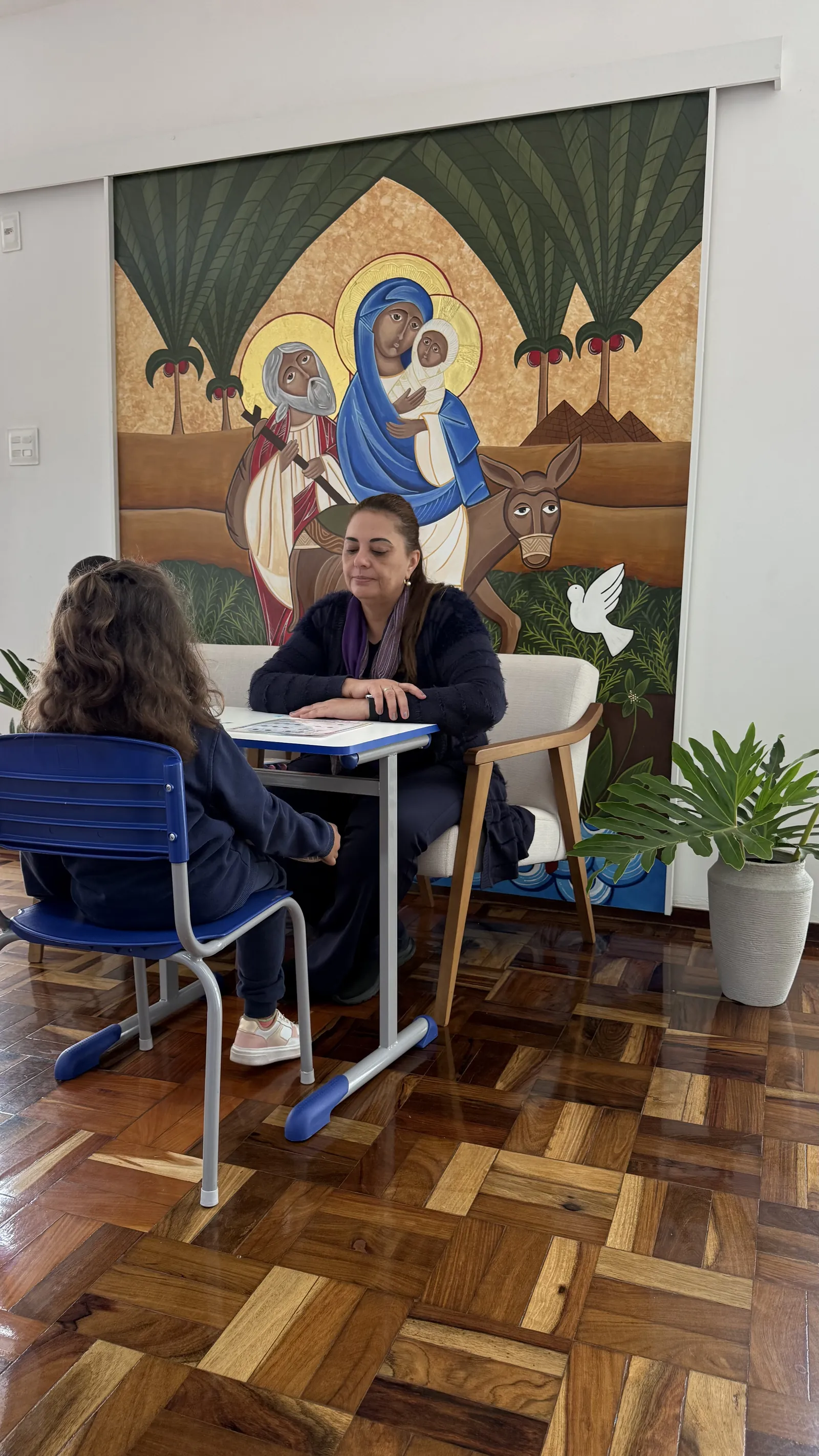 Professora em atendimento individual com aluna, diante de um painel da Sagrada Família