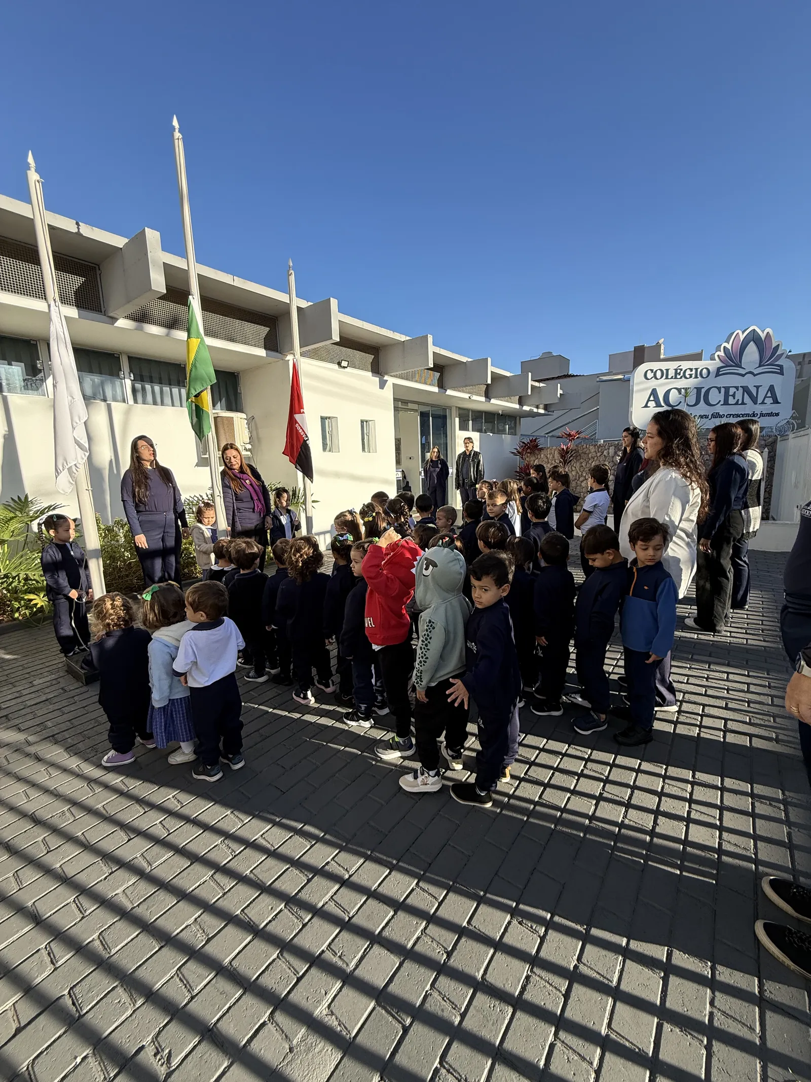 Fachada do Colégio Açucena durante a acolhida, com alunos reunidos e bandeiras hasteadas