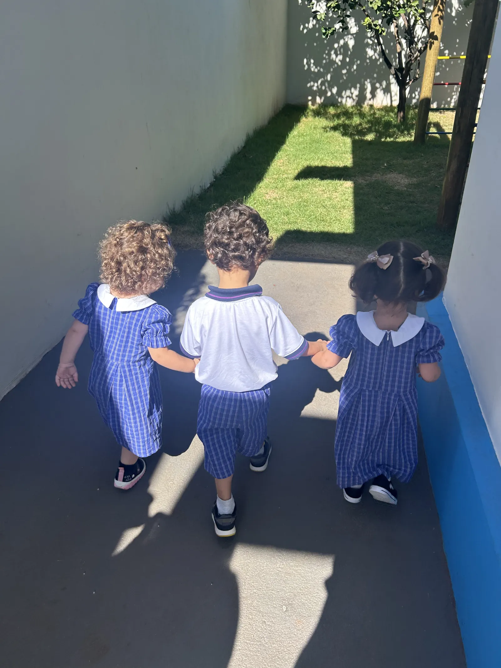 Três crianças pequenas caminhando de mãos dadas pelo colégio