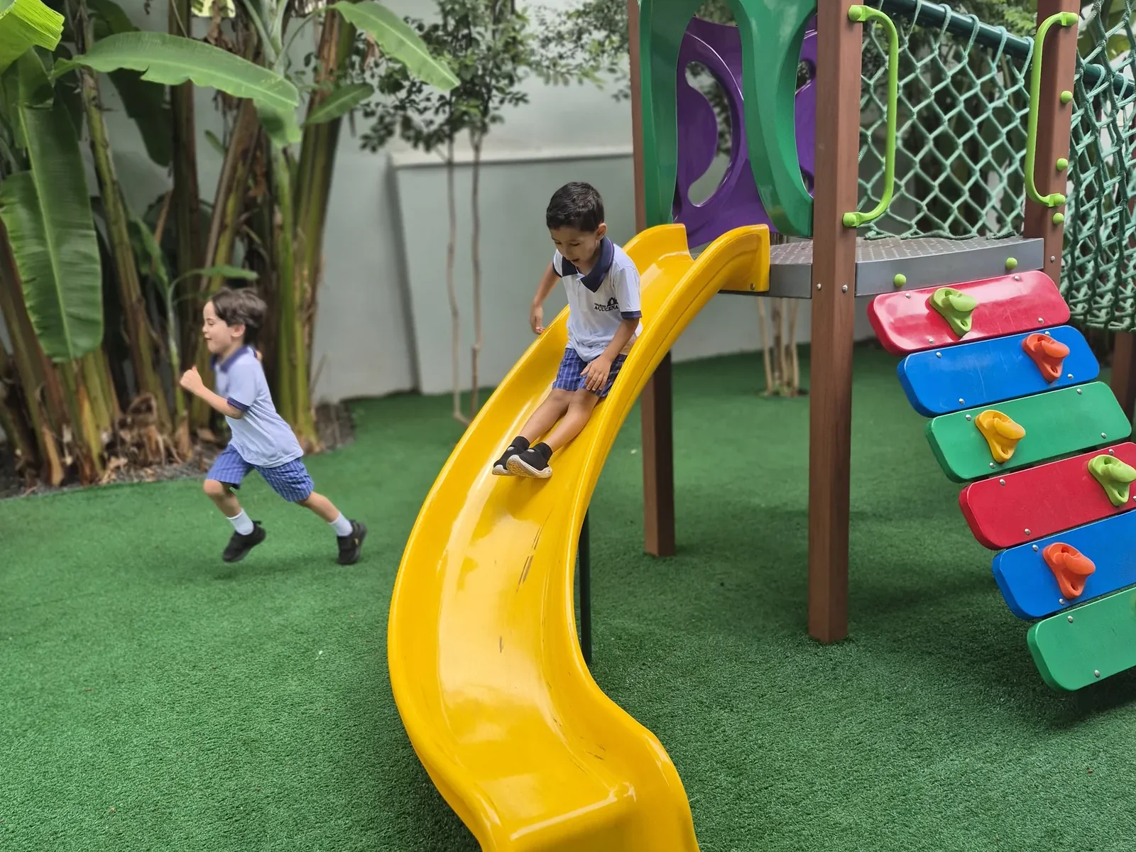 Área verde do colégio com gramado, bananeiras e crianças brincando no escorregador