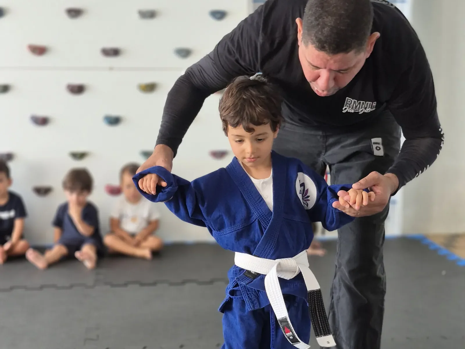 Professor de judô orientando aluno de kimono no tatame, com parede de escalada ao fundo
