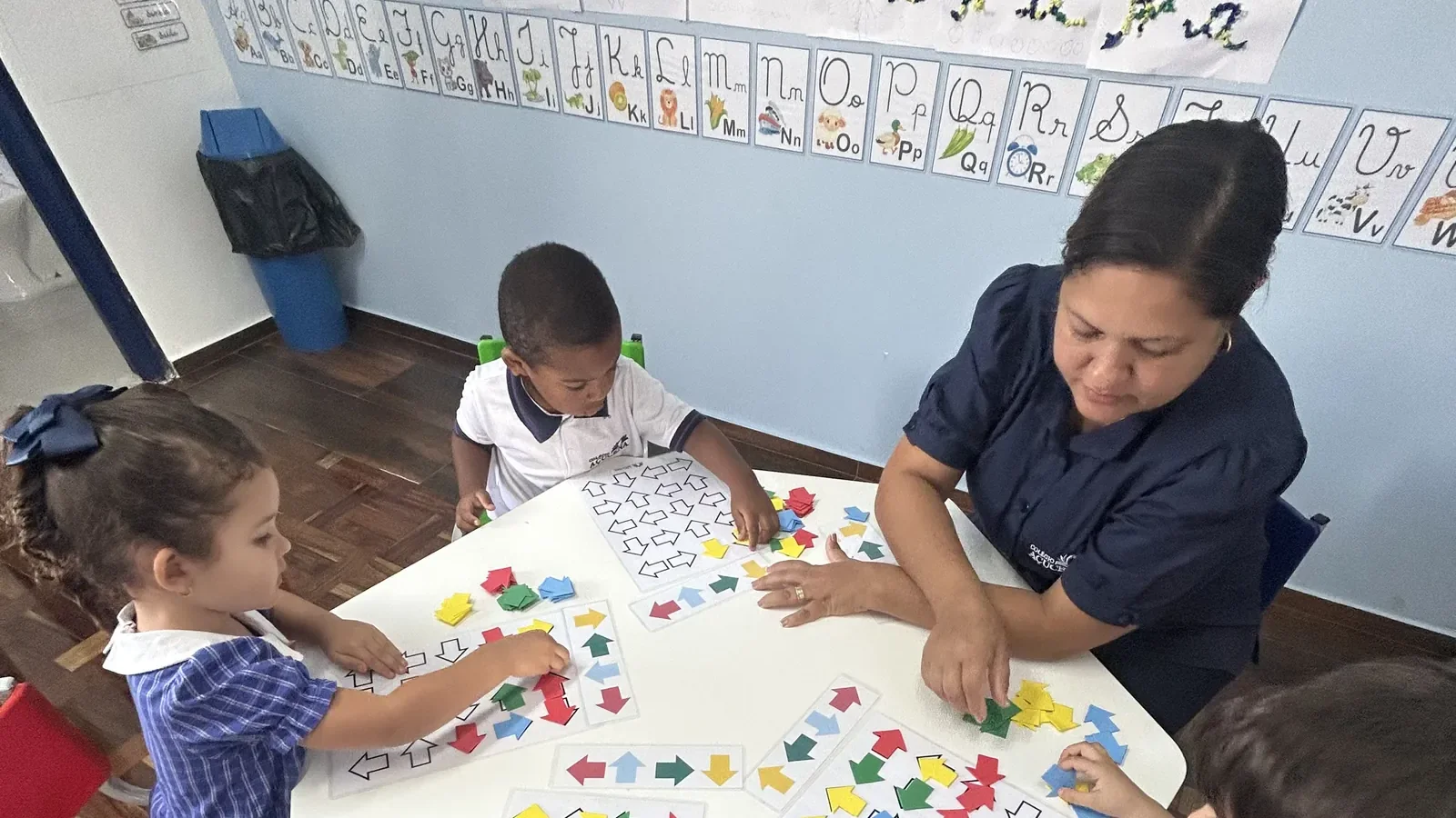 Sala de aula em uso: educadora conduz jogo pedagógico com as crianças, com mural de letras na parede